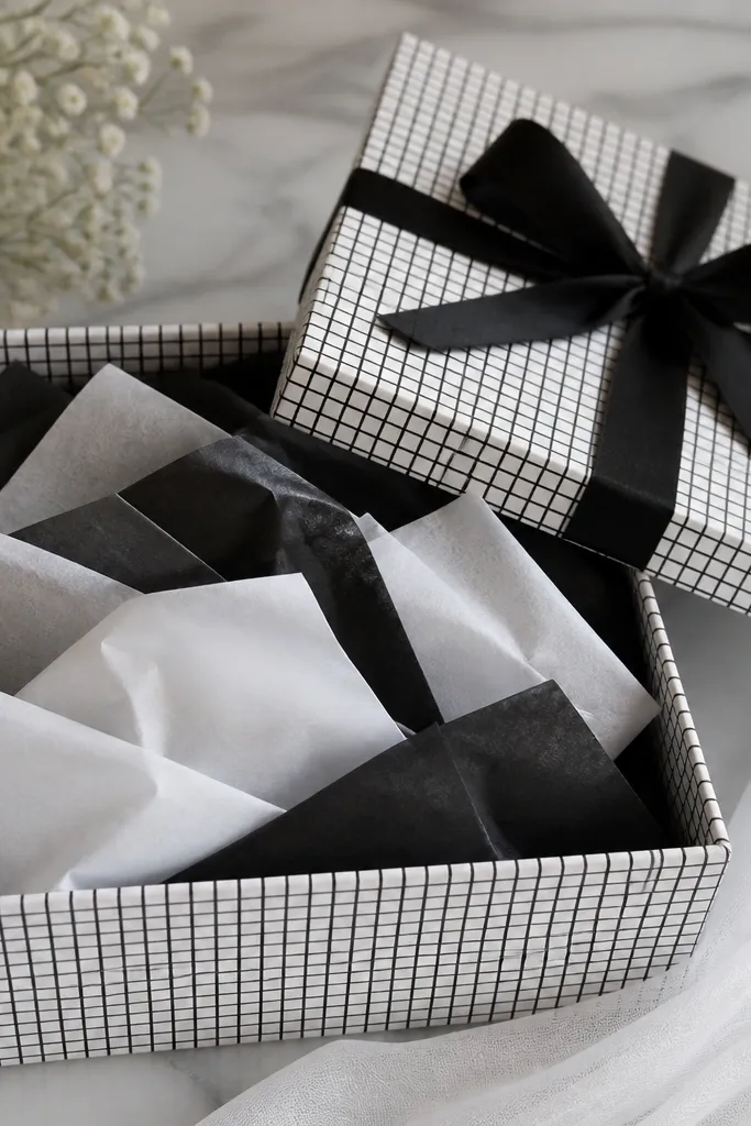 A box with a black-and-white grid pattern on the sides. Inside, black and white tissue paper squares are folded and layered. The lid has a plain black ribbon tied in a neat bow.