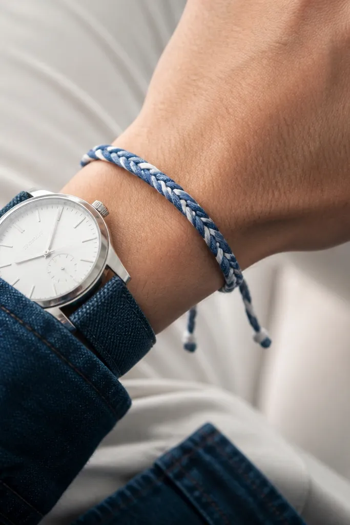 A slim bracelet made from braided denim strips with a white thread accent and a sliding knot closure; it's photographed on a wrist with a watch.