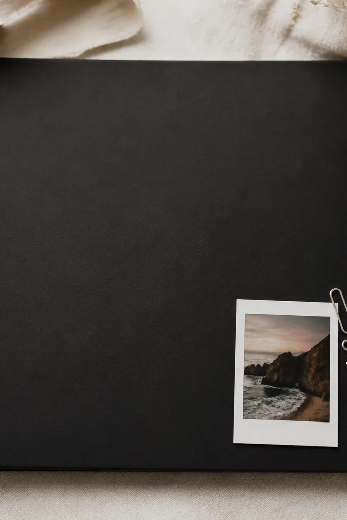 A matte black scrapbook cover with crisp white vinyl lettering centered on the top half. On the bottom right, there's a micro polaroid-style photo with a thin white border. A small silver paperclip charm hangs from a corner.