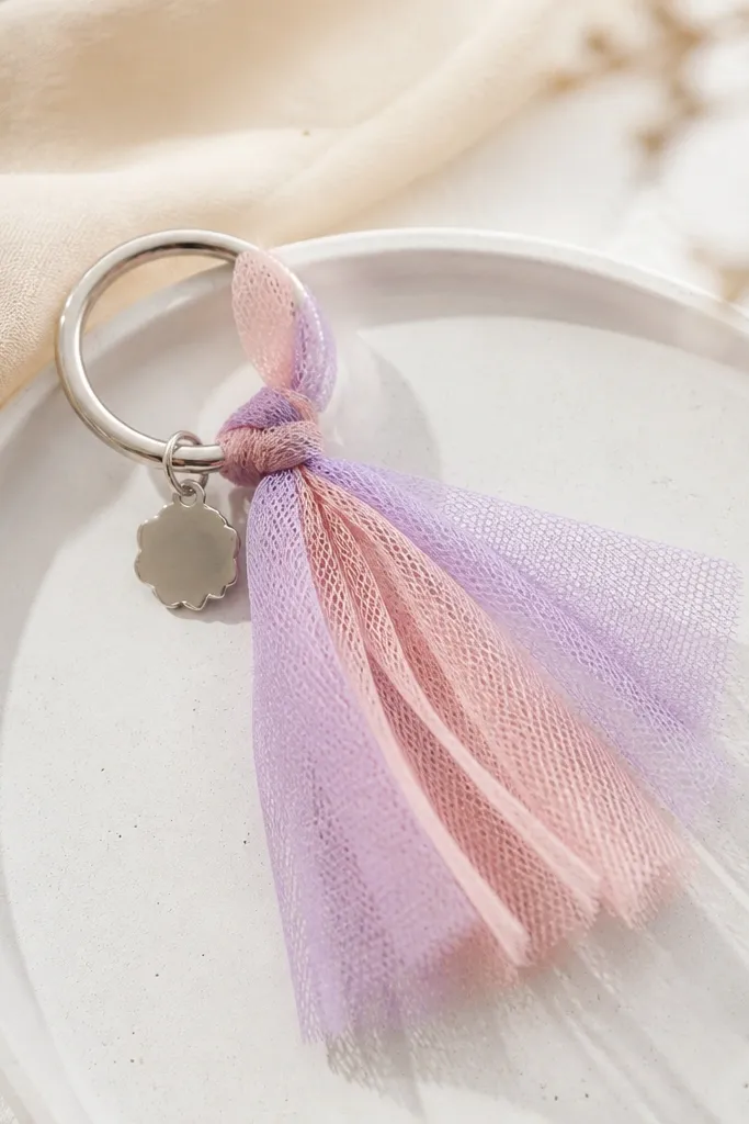 A keychain with a small cluster of tulle strips and a mini charm tag, tied around a metal ring, in pastel pink and lavender.
