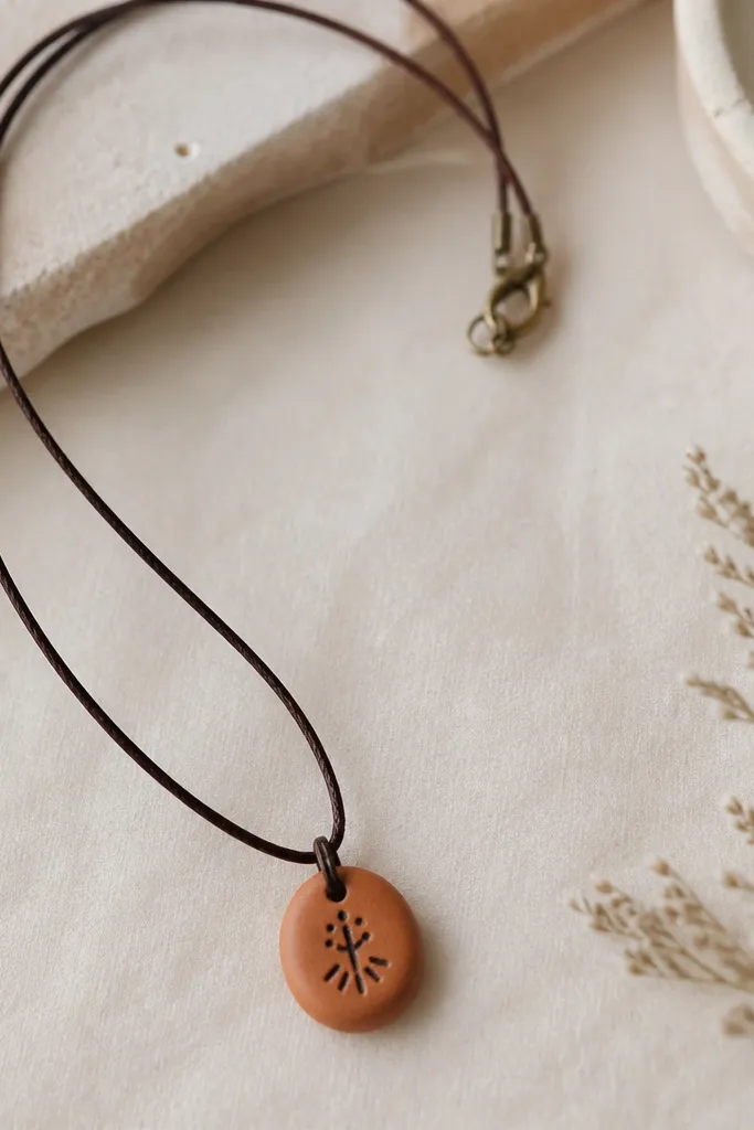 A simple cord necklace with a small stamped clay pendant, light terracotta color, with a black stamped symbol and a thin metal clasp.