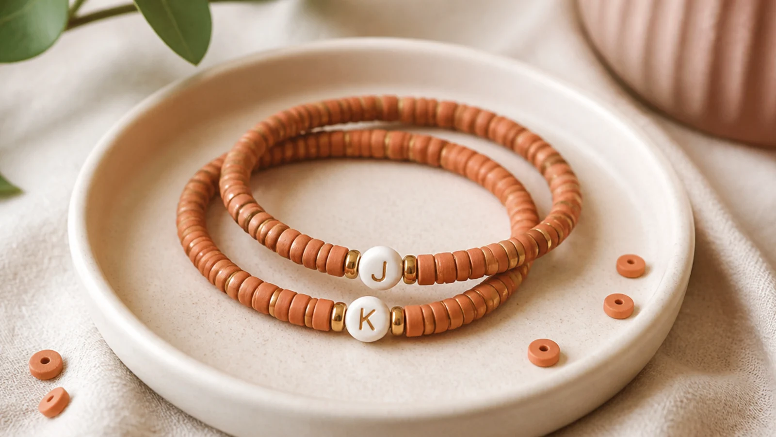 Two clay bead bracelets on a matching base, each showing the other person's initial in a white letter bead.