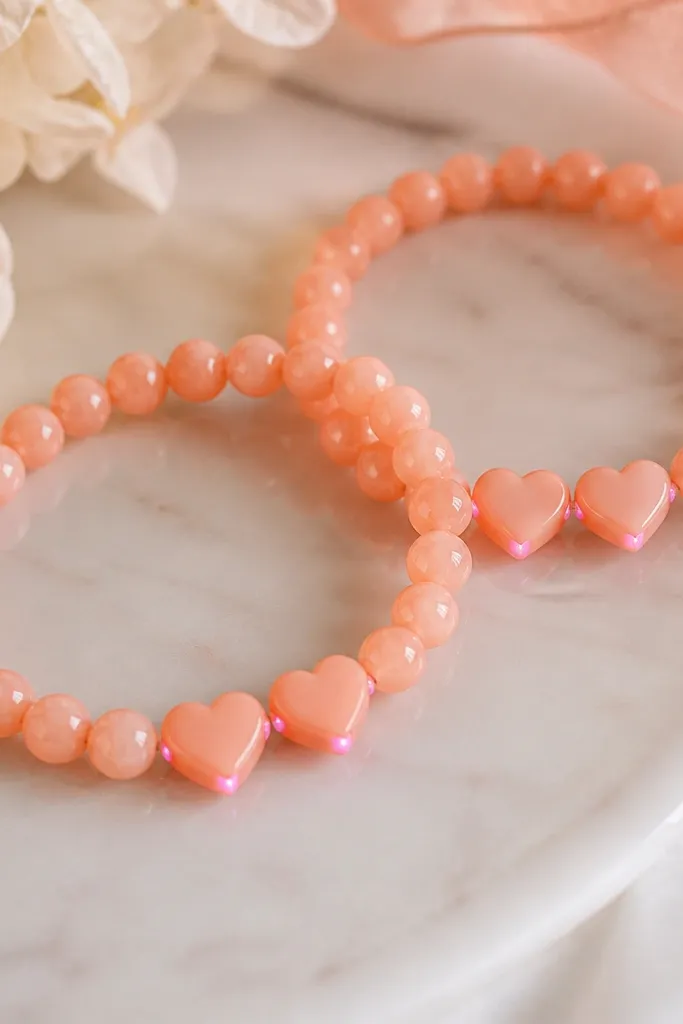 Two matching coral peach bracelets with 8mm beads and two tiny heart-shaped clay pieces on each bracelet. Pink LEDs glow at the heart corners rather than across the whole bracelet. Wire is hidden under peach bead caps.