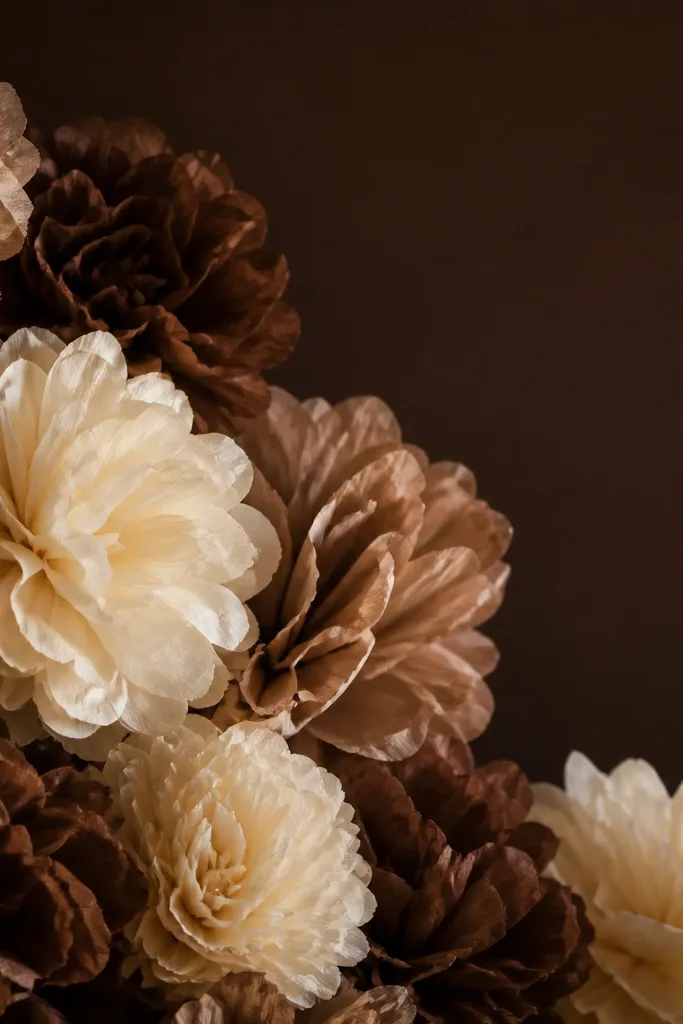 A deep chocolate brown backdrop with cream and mocha tissue paper flowers arranged in clusters. The cream petals pop sharply and look warm under low light.
