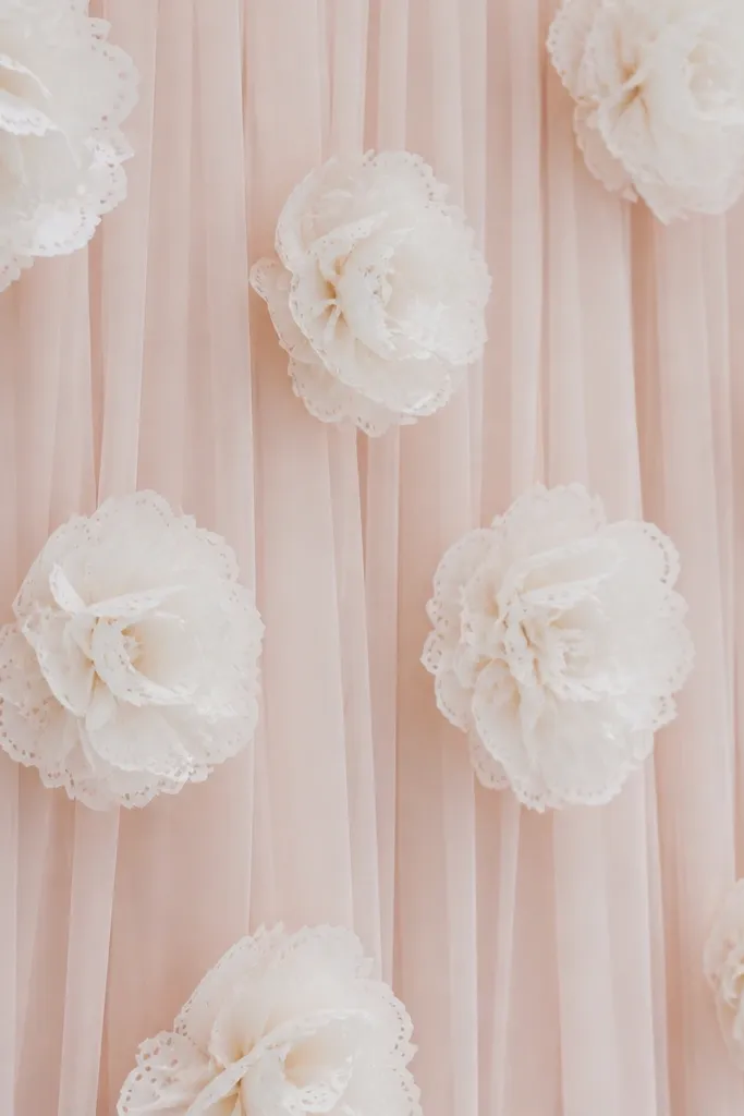 A blush veil fabric backdrop with white tissue flowers that have delicately scalloped petals. The flowers look lace-like and hang slightly away from the background.