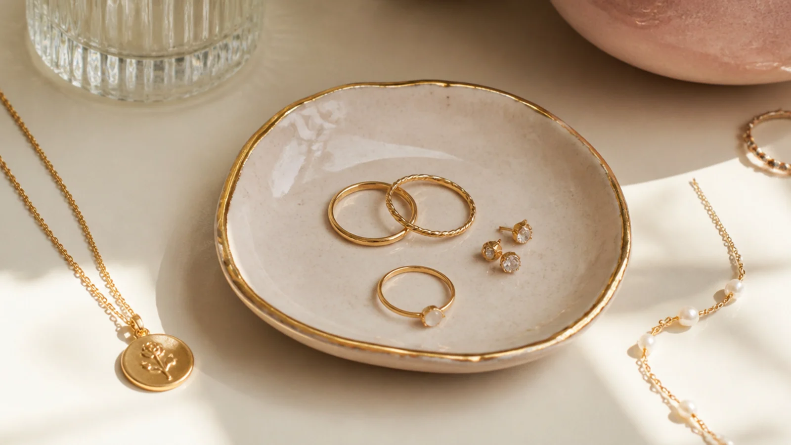 A small handmade air-dry clay dish glazed cream with a gold rim, holding a few rings and stud earrings on a nightstand.