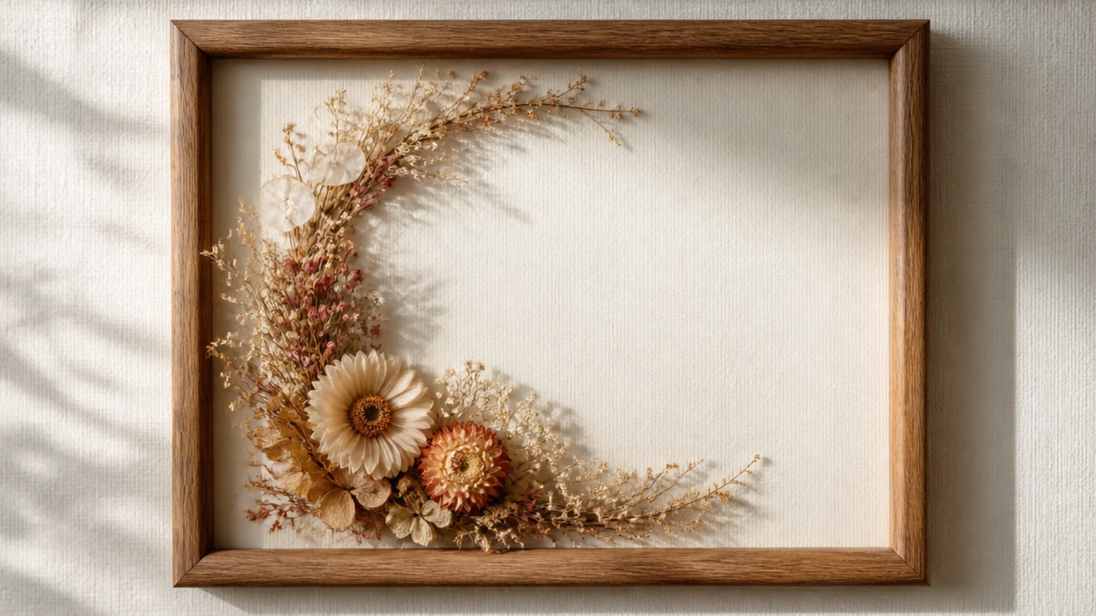 A frame with a half-wreath of dried flowers curving around two corners, framing an open center on cream backing.