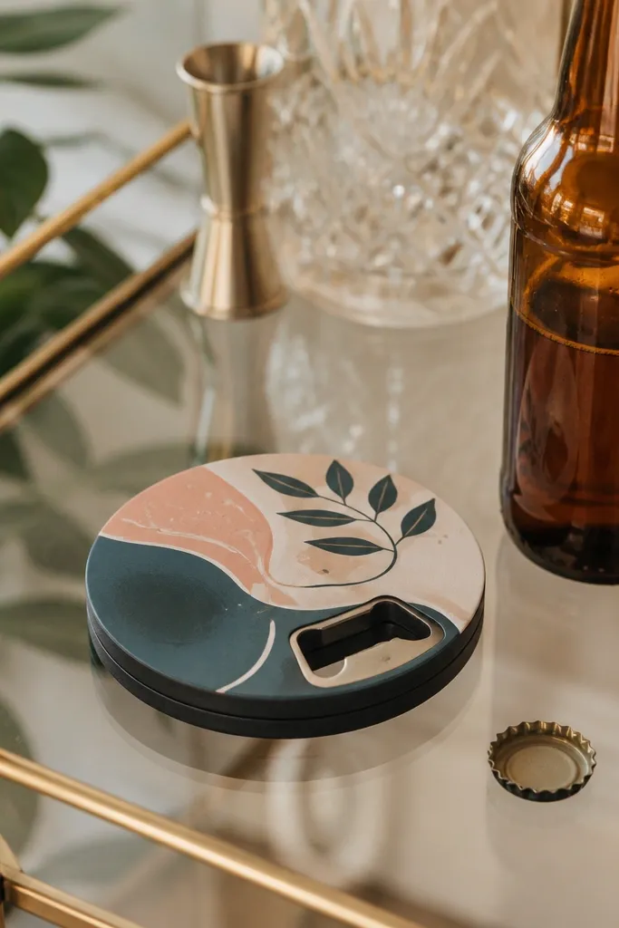 A round coaster with a bottle opener built into the side, customized with a printed label and a small metal opener piece. It's shown next to a bottle on a bar cart.