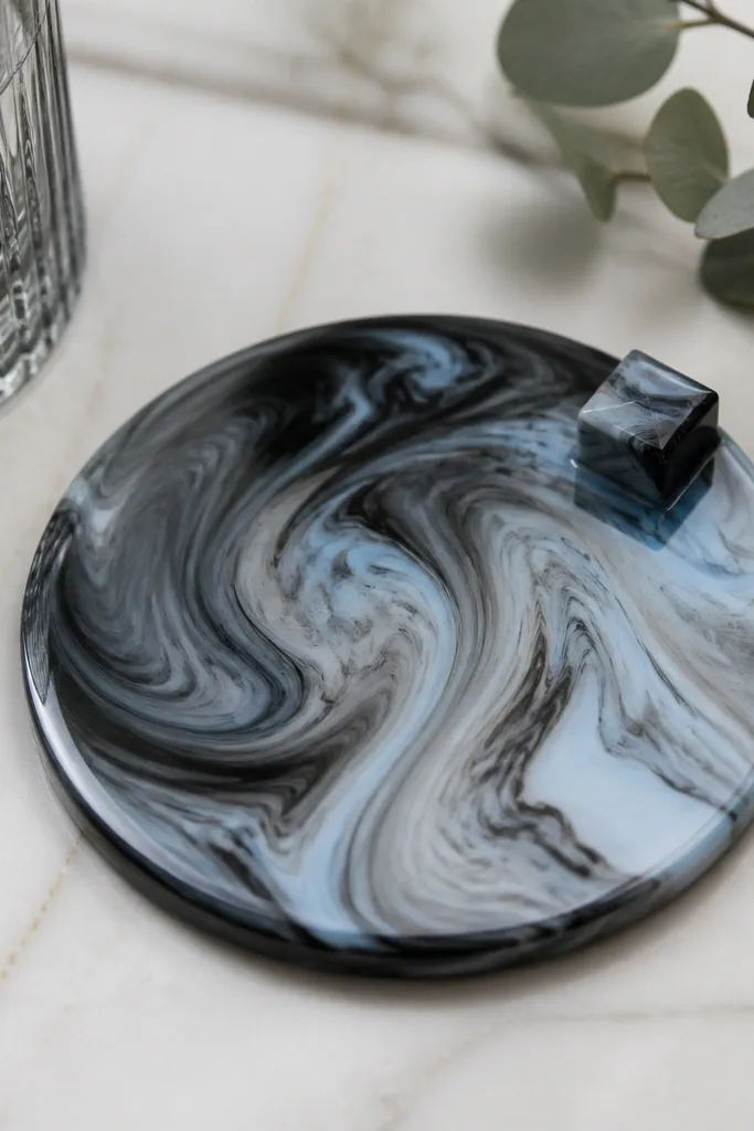A round coaster with a faux marble swirl pattern in black, gray, and pale blue; it has a glossy resin-like finish and a small keycap-shaped accent near the edge.