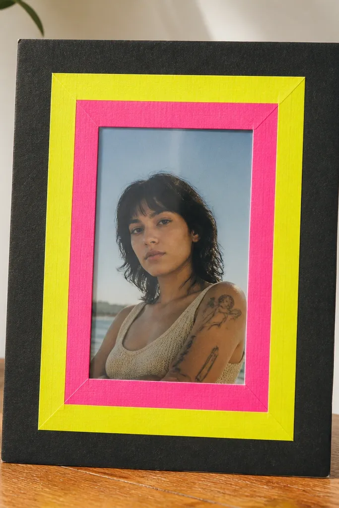 A 5x7 cardboard frame with a bright neon yellow and pink tape border. The tape forms a thick outline around the window, and the photo sits behind clear plastic. The outer cardboard is painted black.