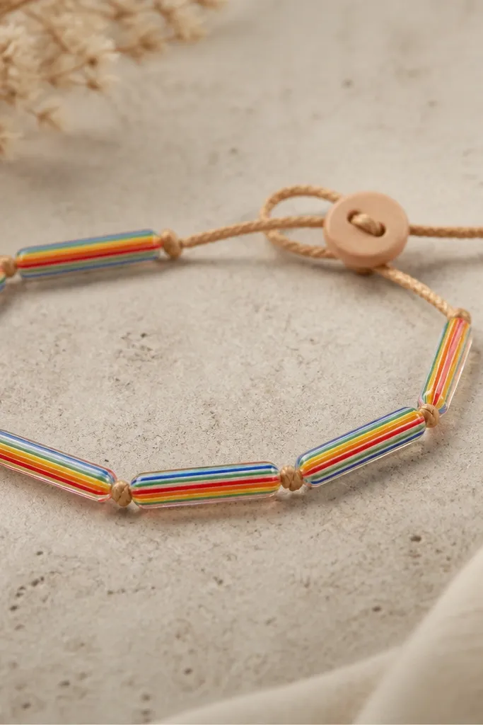 A bracelet with elongated tube beads. Inside each tube, thin rainbow stripes run lengthwise like candy. The bracelet closes with a small light wood slider button on a braided cord.