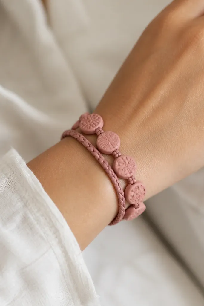 A wrap-style bracelet with two strands of braided cord crossing the wrist. Flat clay medallions in dusty rose are attached along one side, each medallion has a subtle carved pattern.