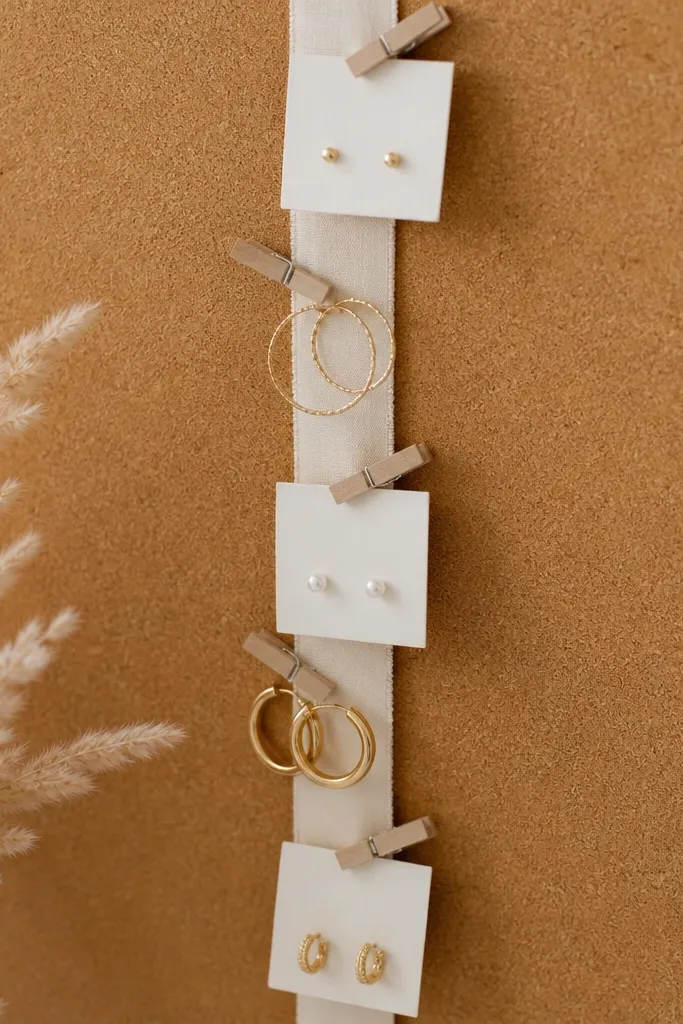 A vertical ribbon strip pinned to a cork board. Clothespins clip small earring cards and a few hoop earrings, with a soft neutral color palette.