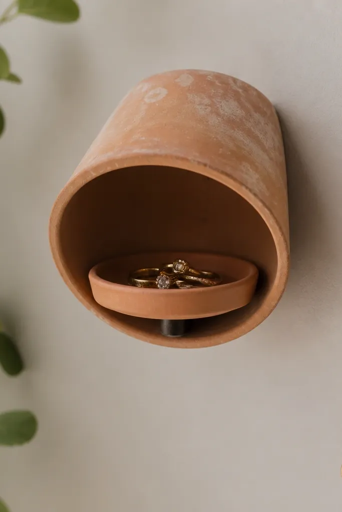 A small shallow terracotta plant pot mounted upside down on a wall with a ring dish inside. Rings sit in the pot's center, and a small hook holds one necklace nearby.