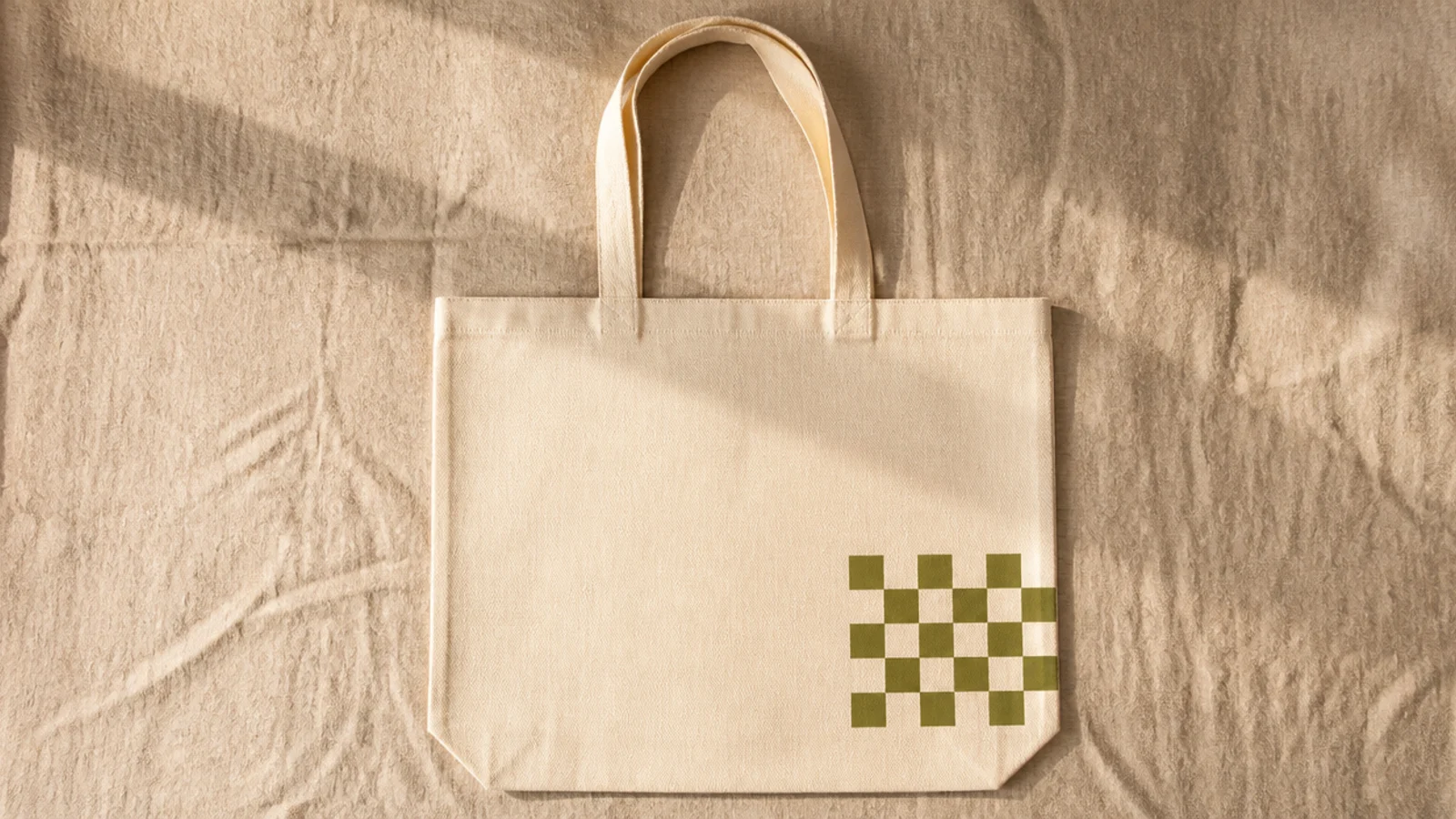 A cream canvas tote with a small block of olive-and-cream checkerboard squares in the lower right corner, the rest plain.
