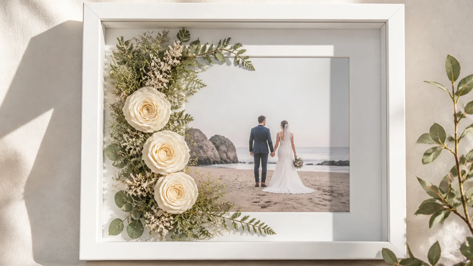 An elegant white frame with preserved white roses and greenery along one side around a wedding photo.