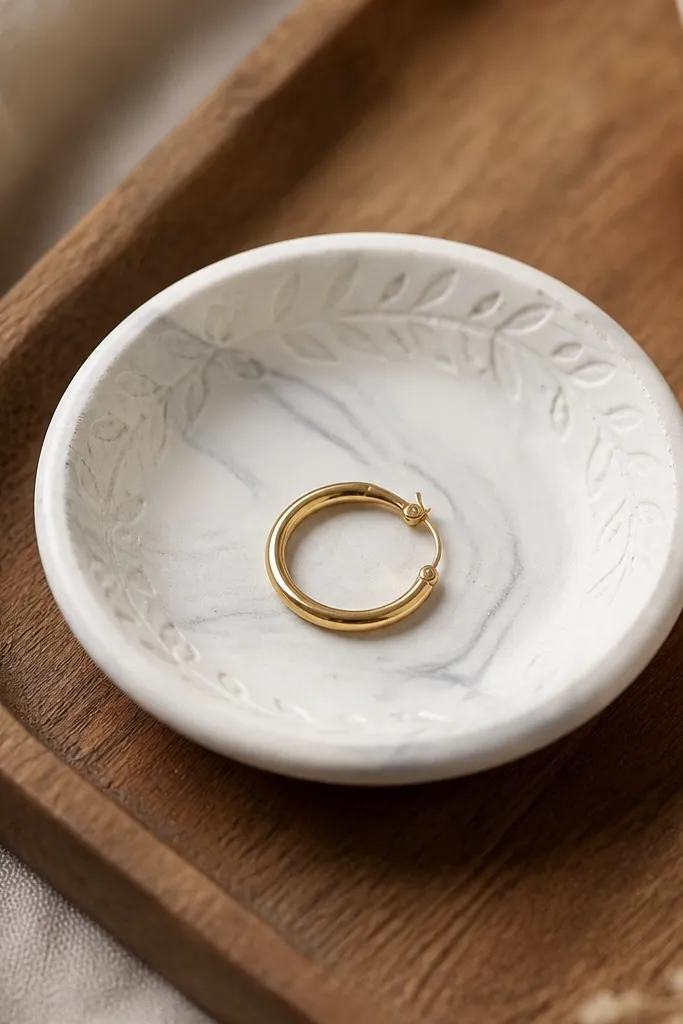 A small round ring dish made of white clay with subtle gray marbling swirls, embossed with a faint leaf pattern around the rim. It sits on a wooden tray, with one gold hoop earring resting inside.