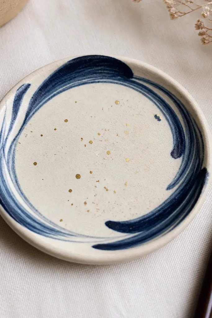 A small round ceramic dish the size of a coaster. The dish has a matte cream base with a hand-painted navy swirl around the rim. Tiny gold speckles scatter across the center like confetti. A matching mini brush is visible on the side for scale.