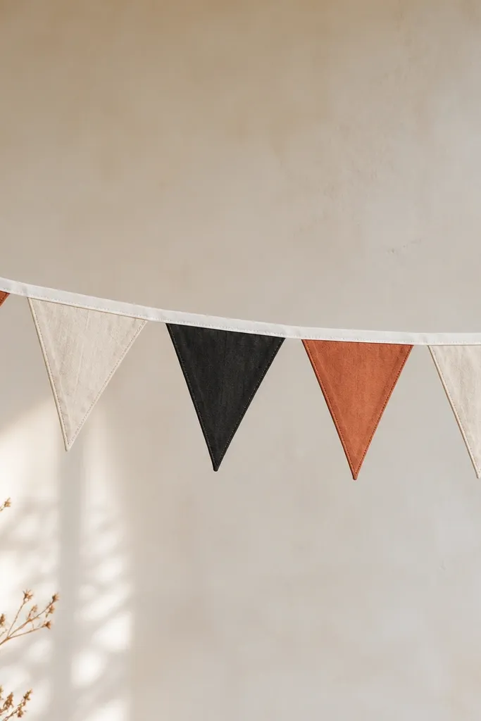 A string banner made of small triangle pennants in three fabrics: cream linen, black cotton, and a muted rust-orange. The triangles hang from a thin white twill tape line, evenly spaced.