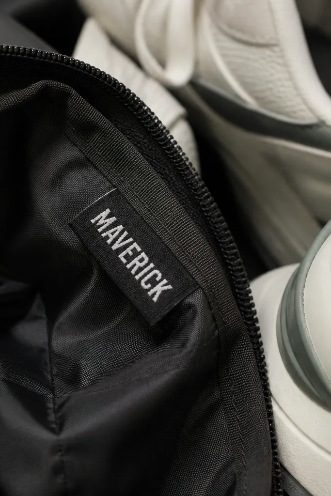 A black fabric laundry tag sewn to the inside seam of a gym bag, with white heat-transfer letters reading a nickname, photographed next to a pair of sneakers.