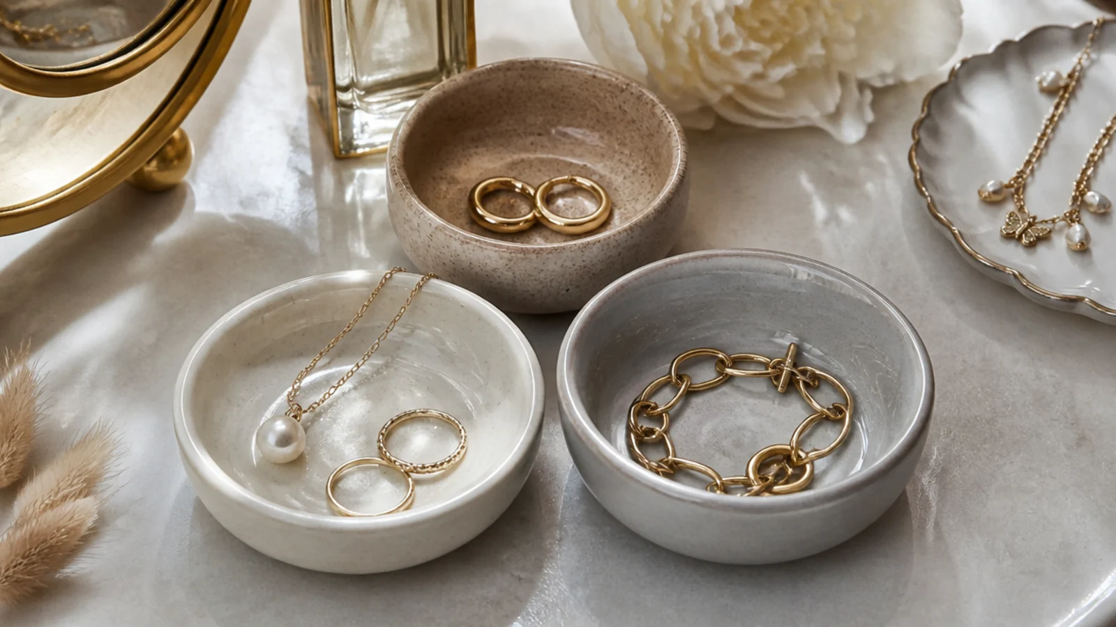 Three small ceramic bowls in cream, sand, and pale grey grouped on a vanity, each holding one type of jewelry.