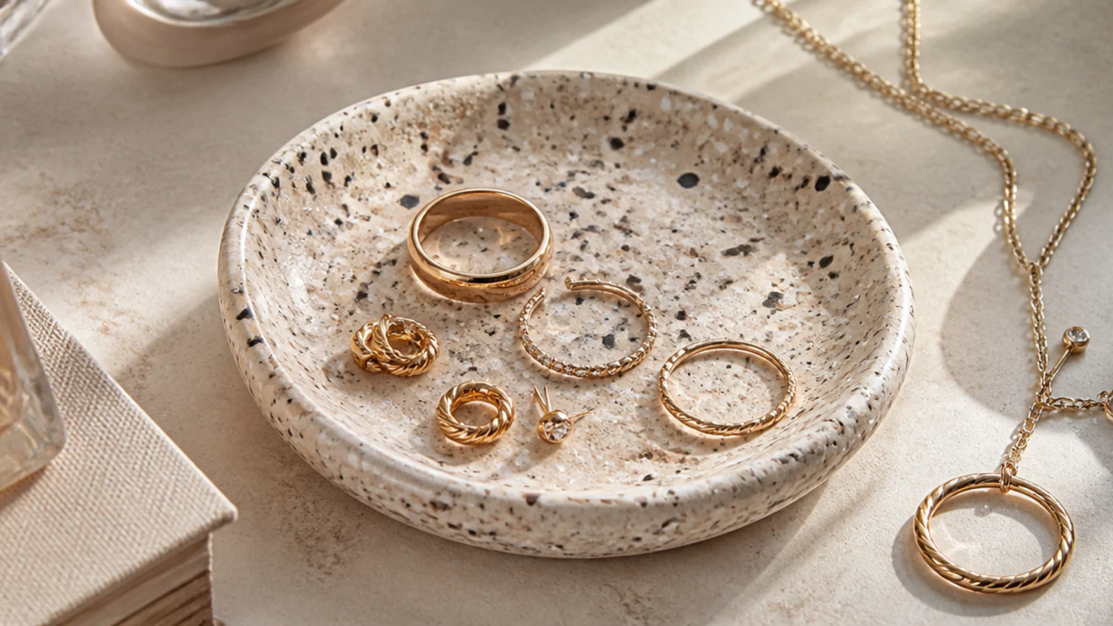 A shallow handmade clay dish with a pale terrazzo speckle pattern holding rings and small earrings on a nightstand.