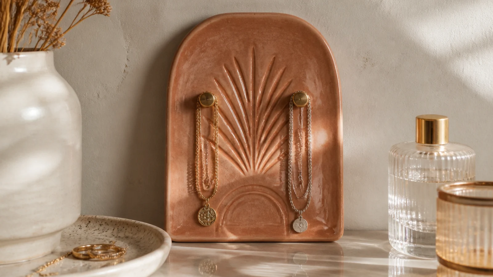 A small glazed clay wall plaque with two embedded brass hooks, holding a couple of necklaces against the wall.