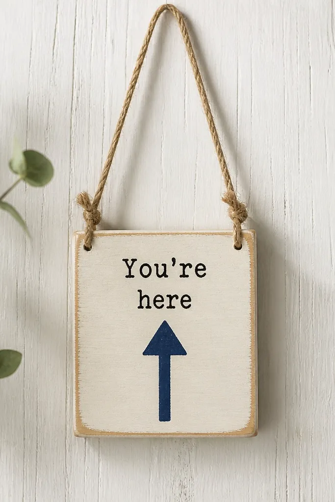 A small wooden wall hanger painted in soft cream with a navy painted arrow and the words "You're here" in simple black letters. A single twine loop hangs from the top, and the wood grain still shows around the edges.