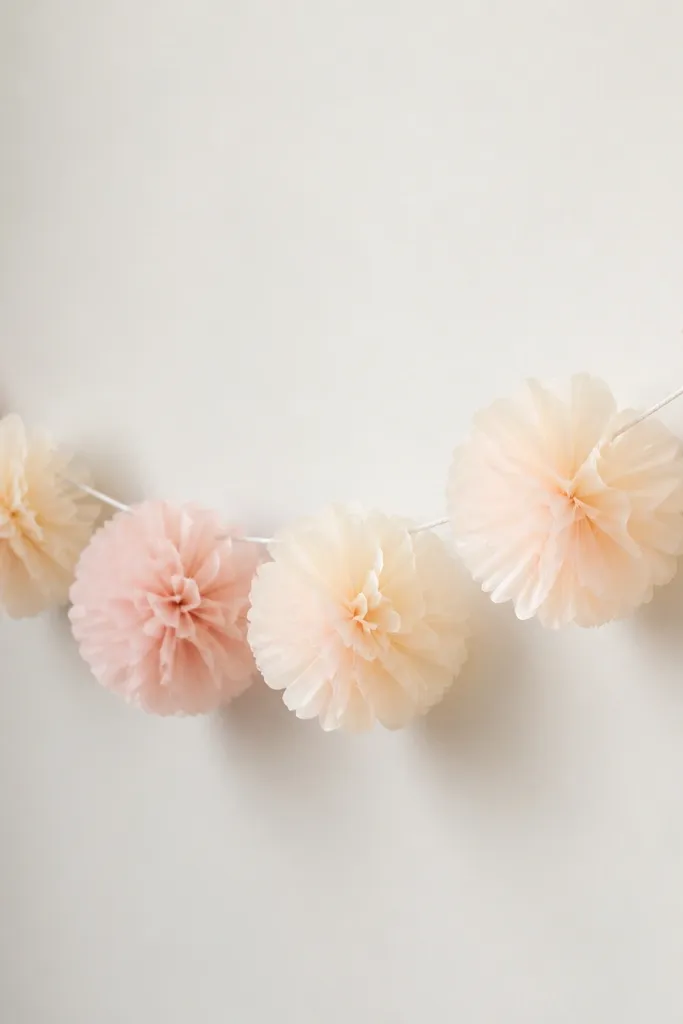A garland made of tissue paper pom-pom style flowers in soft cream and pale blush, strung along a thin white cord. Each flower has layered fan petals with a slightly ruffled edge, spaced evenly across a light wall.
