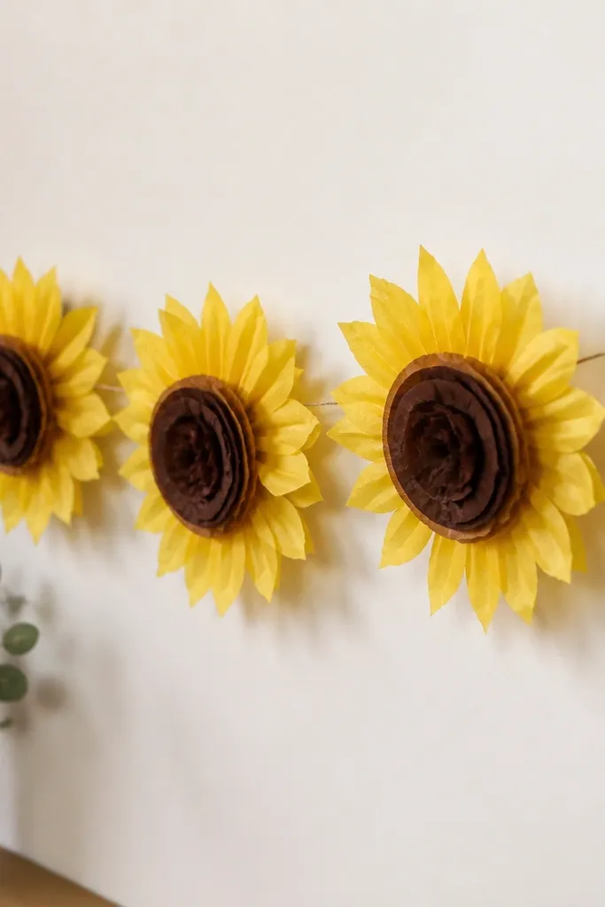 Tissue paper sunflower-style garland with bright yellow petals and dark brown centers. The centers are layered tissue circles, and the garland runs across a wall.