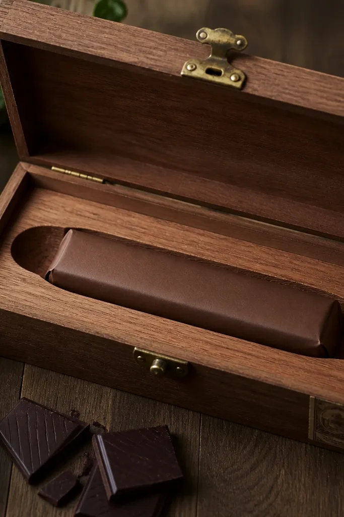 A cigar-shaped insert cut from cedar wood with a dark chocolate bar wrapped in brown paper; the box has a brass clasp and a small stamped label.
