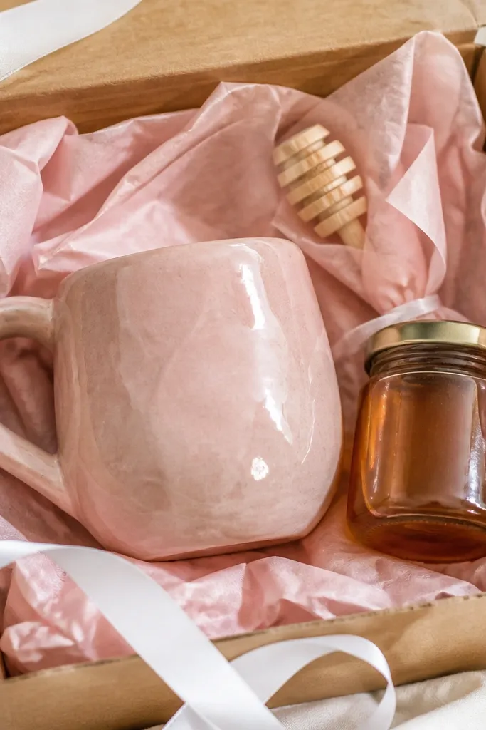 A blush-pink glazed handmade mug with a glossy finish. A jar of honey sits beside it, with a small wooden honey dipper wrapped in tissue. The box has soft pink tissue and a white ribbon.
