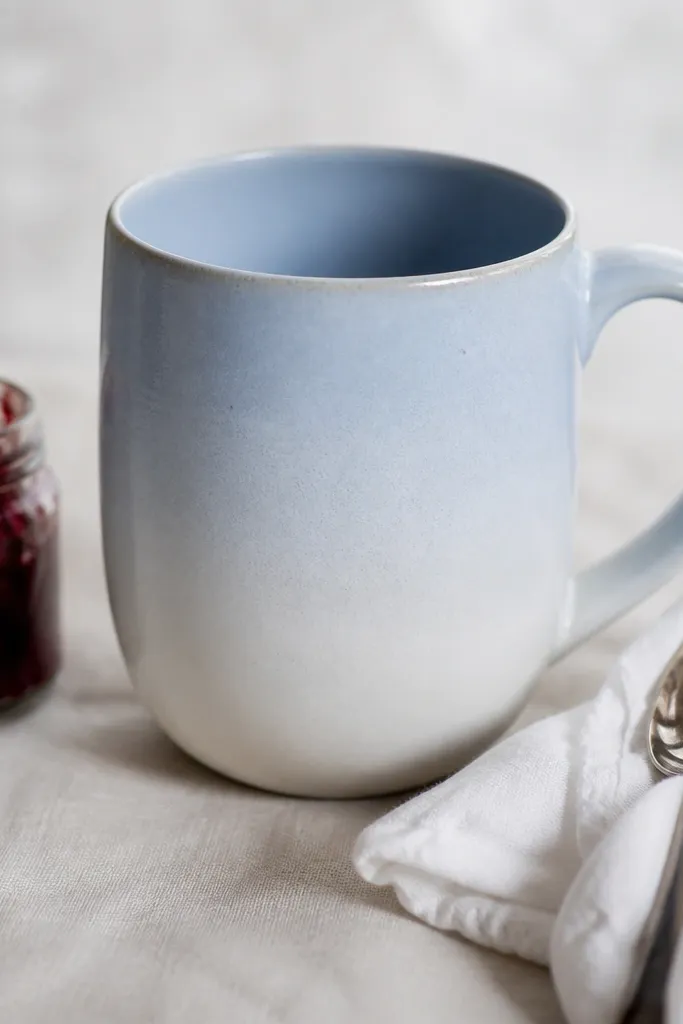 A powder blue mug with a soft ombre fade, shown with a tiny jar of berry jam and a spoon wrapped in white cloth.