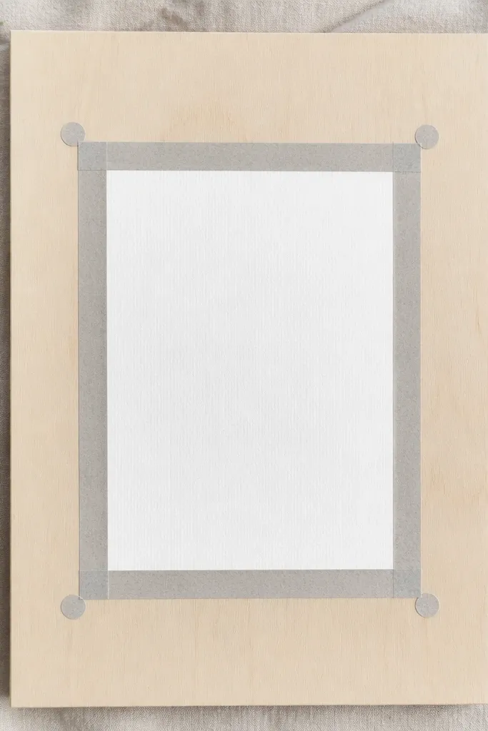A 12x16 board with light wood. Pale gray washi tape forms a rectangle frame around a white card. Two small dots of tape sit at the corners for balance.