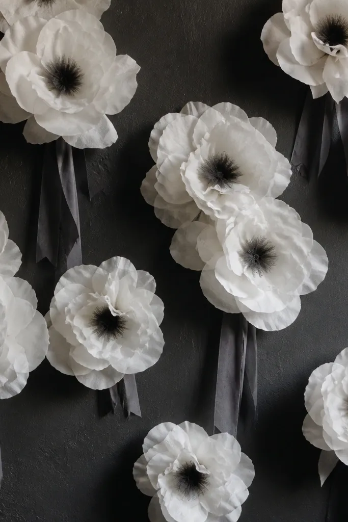 White tissue paper flowers with matte outer petals are scattered across a dark wall. Each cluster has a few charcoal-gray ribbon strips tied around the stems, and the centers are darkened with a charcoal tint so the blooms look high-contrast.