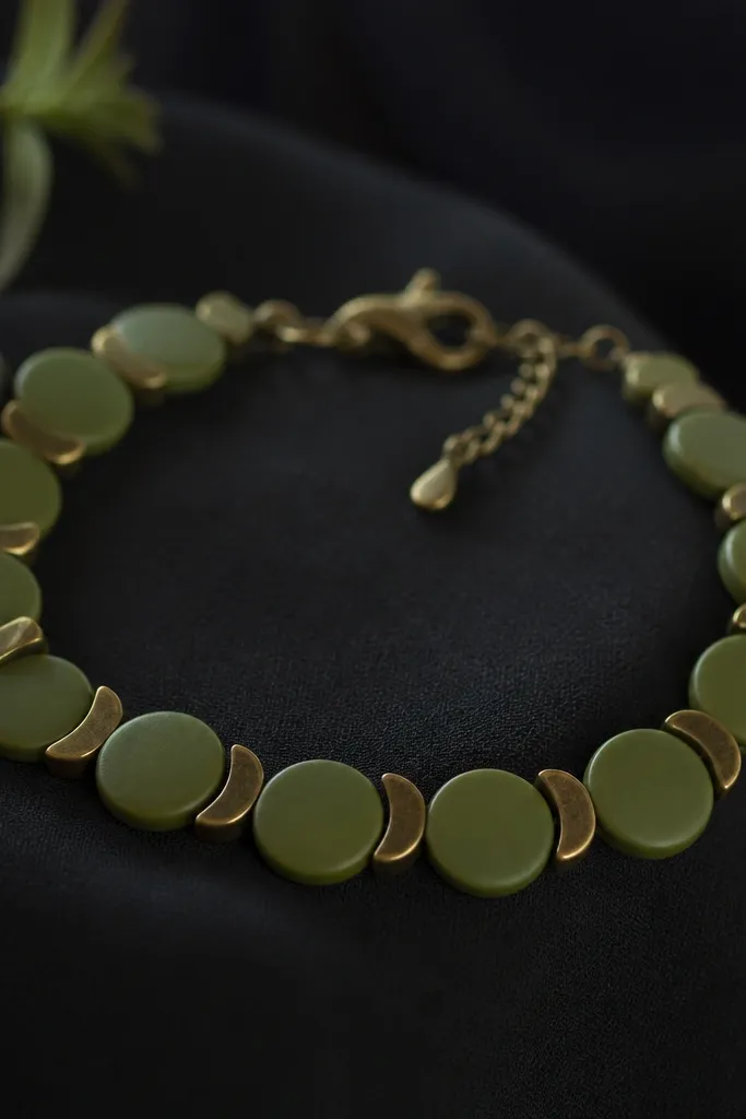 A bracelet made of flat moss green clay coin beads (9mm) alternating with blackened brass half-moon spacers, photographed against dark fabric.