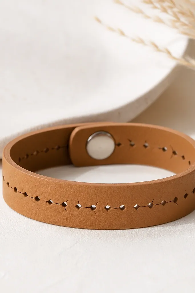 A tan leatherette bracelet with a repeating perforated pattern in a straight line. A small silver snap clasp closes the bracelet. It looks smooth and structured.
