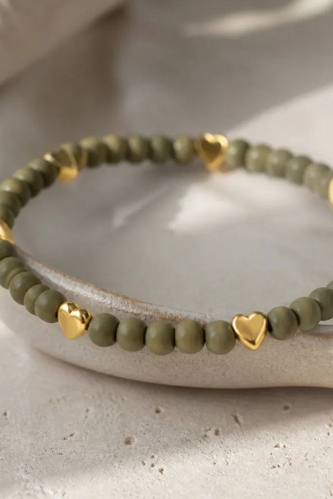 An olive clay bead bracelet with small gold heart charm pieces spaced at intervals. The hearts sit between beads and catch light.