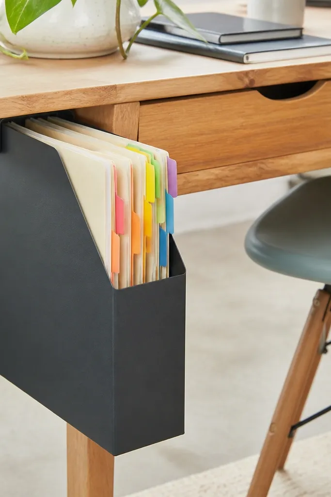 A slim magazine file hangs or sits under the desk. It holds folders standing upright. DIY label tabs in bright colors are attached to each folder edge.