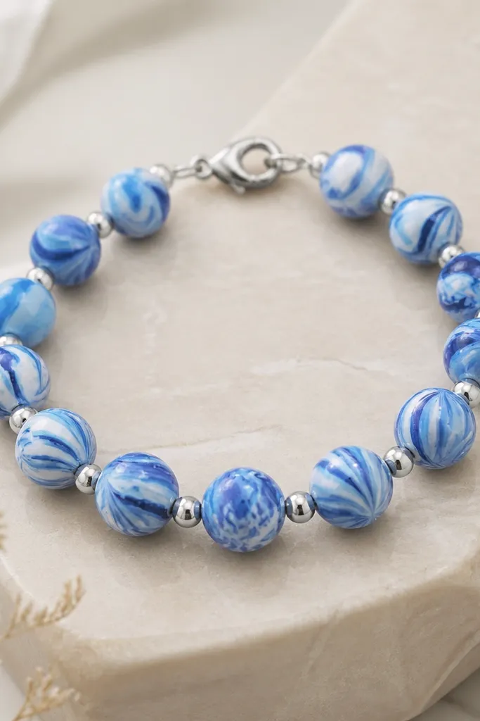 A bracelet showing blue marble clay beads in varying swirls. Between each bead is a single silver divider bead, and the clasp is a small silver lobster clasp.