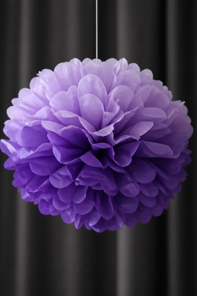 A hanging tissue flower pom in purple ombre, with the top lighter and bottom darker. It's suspended from a string and photographed against a dark curtain.