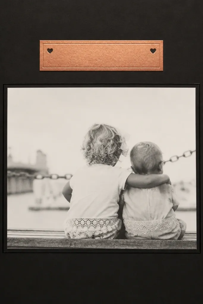 A page with a deep black cardstock background. Centered is a matte white photo with a thin black frame. Above it sits a small rose gold foil label with a short quote, plus two tiny heart icons in the corners.