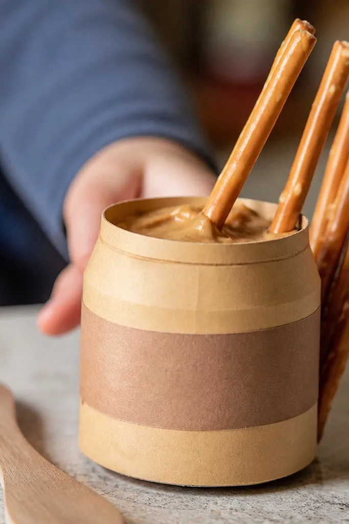 Paper jar of peanut butter with small pretzel sticks dipping in