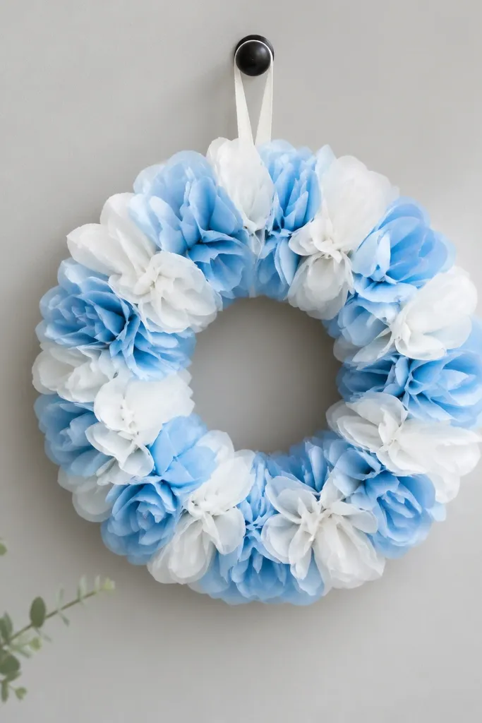 A small circular wreath made of layered tissue petals in white and light blue. It hangs on a wall hook and sits flat with a clean outer rim.