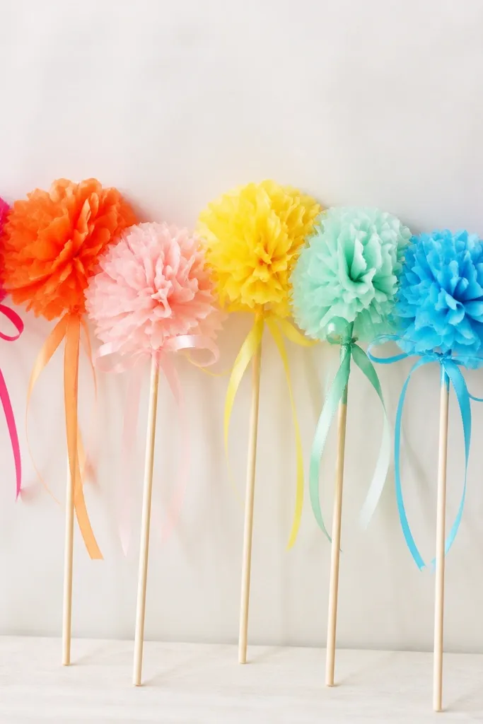 A row of small tissue flower wands on sticks, each bloom around 5 inches wide, in bright colors. The flowers are attached to wooden skewers and tied with ribbon.