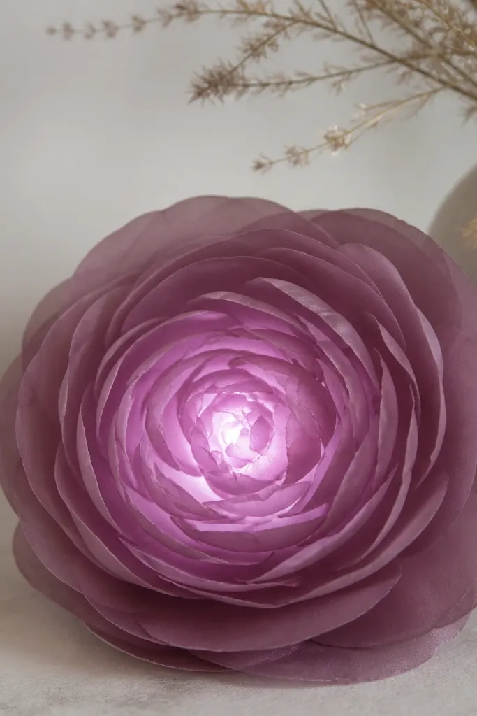 A mauve ranunculus-style flower with tight layered petals, showing a soft mauve glow from a hidden LED behind a tissue diffuser disc.