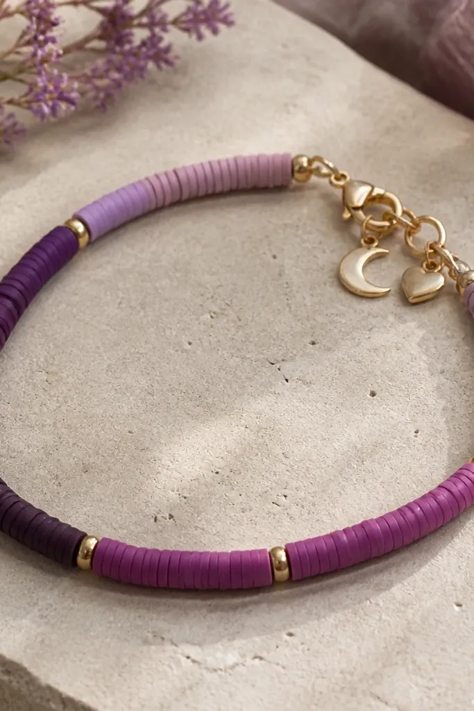 A plum ombre bracelet with small purple clay beads in gradient shades and two tiny metal charms near the clasp; the charms are a moon and a heart.