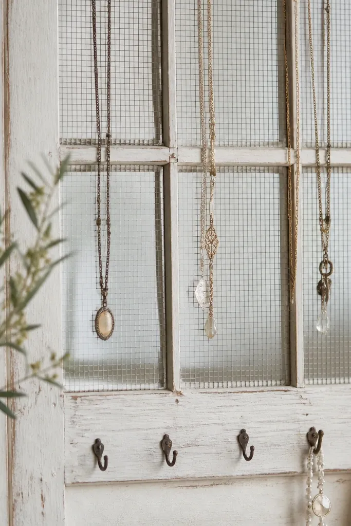 An old wooden window frame painted off-white, with fine metal mesh stretched across panes; necklaces hang over mesh lines and small hooks sit on the lower rail.